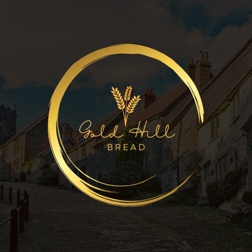 Logo for Gold Hill Bread featuring a gold circular design with wheat stalks inside, set against a background of a quaint village street with cottages and a cobblestone path.