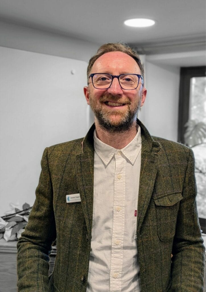 A man with glasses and a beard smiles, standing indoors. He wears a tweed jacket over a button-down shirt. A name badge is on his jacket. A window and desk piled with papers are visible in the background. A man with glasses and a beard smiles, standing indoors. He wears a tweed jacket over a button-down shirt. A name badge is on his jacket. A window and desk piled with papers are visible in the background.