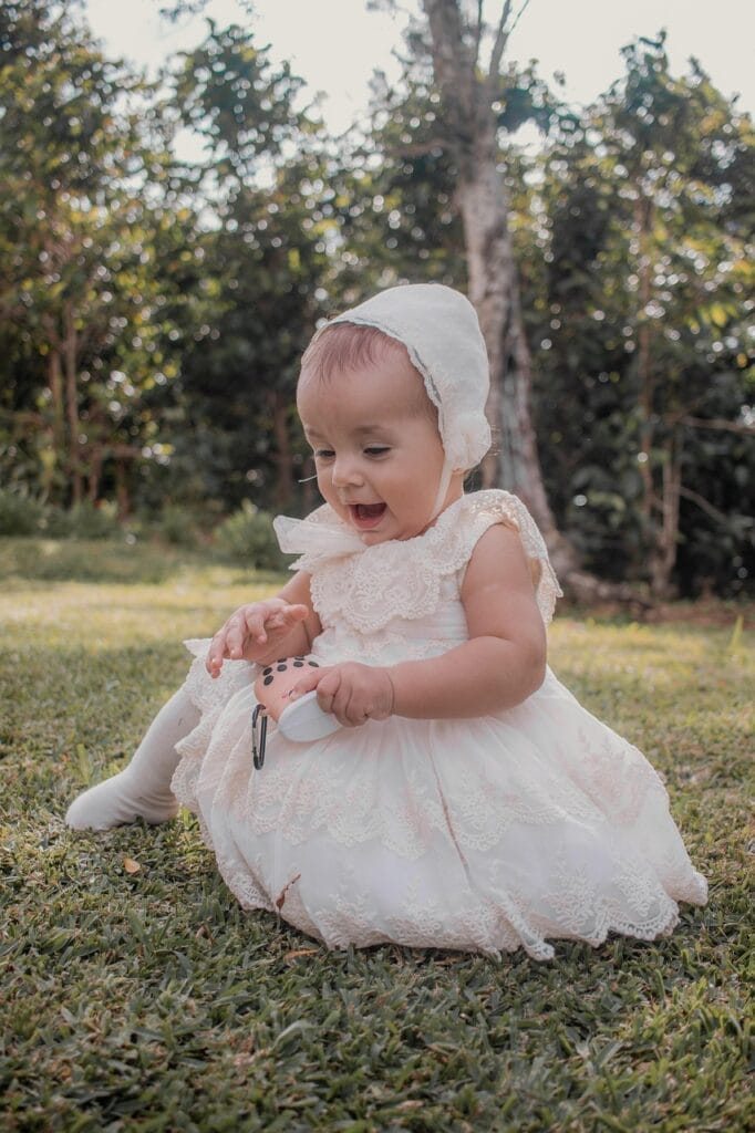 A baby wearing a white lace dress and bonnet sits on the grass, smiling while playing with an object. There are trees and bushes in the background, suggesting a sunny day in a garden or park. baby, infant, christening, baptism, little girl, portrait, baby, christening, christening, christening, christening, christening, baptism, baptism, baptism, baptism, little girl