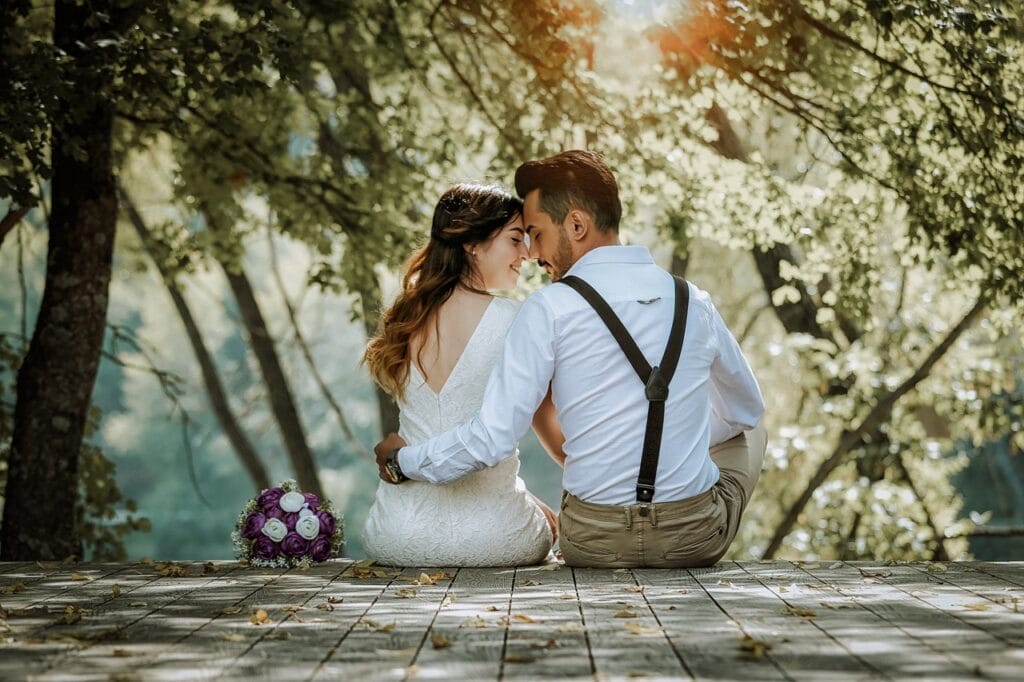 A couple in wedding attire sits on a wooden deck in a forest, nestled together and looking at each other. A bouquet of purple and white flowers rests beside them. Sunlight filters through the trees, creating a serene and romantic setting. happy valentines day, couple, wedding, marriage, bridal, double, nature, love, people, man, woman, romance, happy, romantic, dress, married, male, big, girl, happiness, celebration, ceremony, wife, bouquet, grass