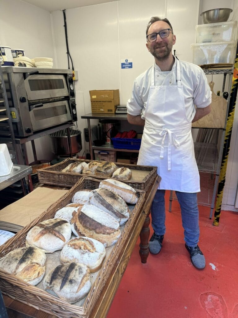 A baker in an apron and glasses stands smiling in a kitchen next to a table with baskets of freshly baked bread. Ovens and kitchen equipment are visible in the background.