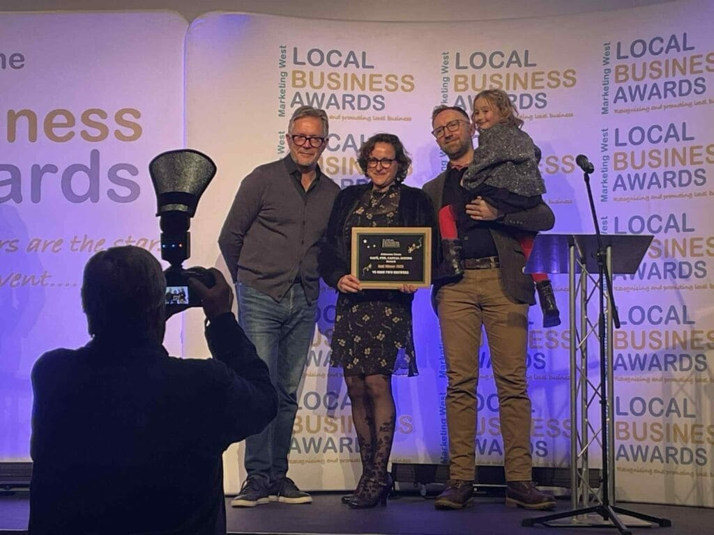 Four people stand onstage at the Local Business Awards, one holding a plaque and another holding a child. A photographer in the foreground takes their picture in front of a branded backdrop.