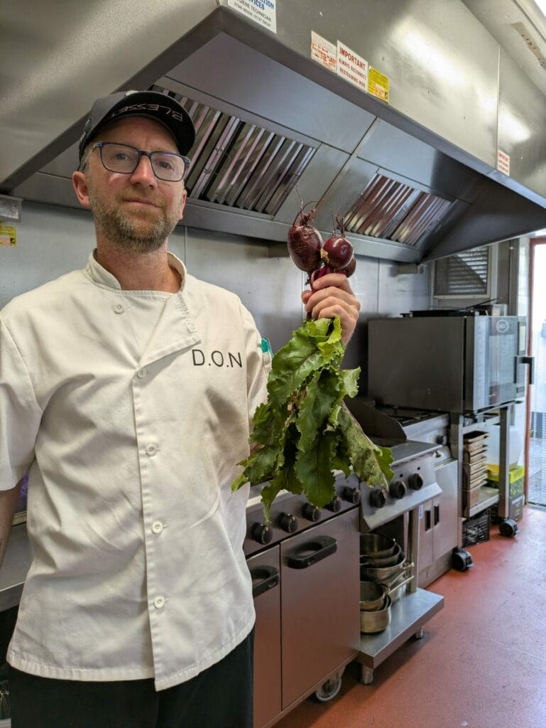 A chef in a white coat and black cap holds a bunch of fresh beetroot in a commercial kitchen, with cookers and utensils visible in the background.
