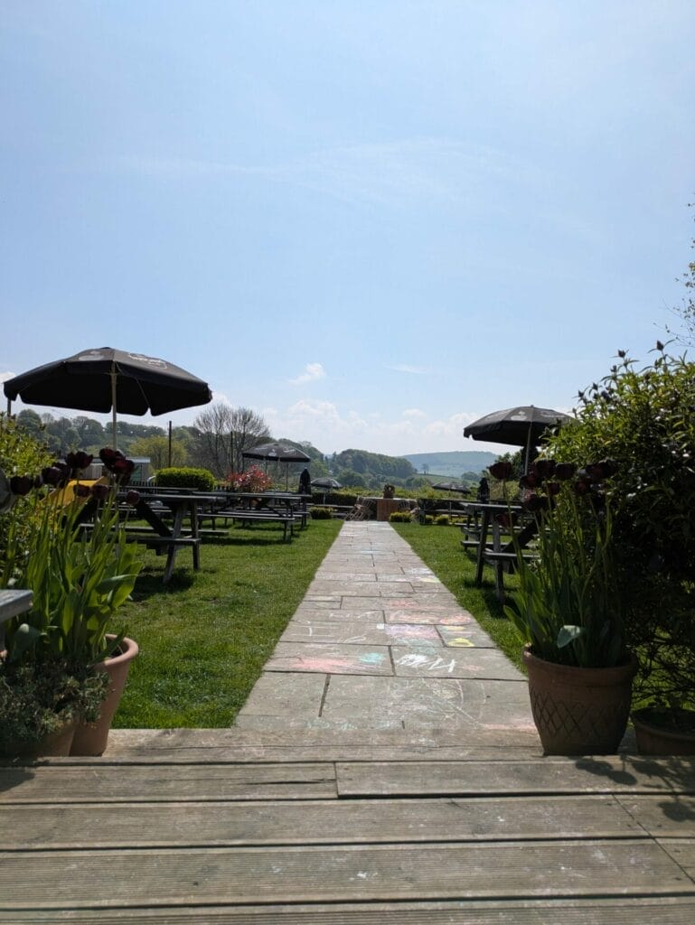 A stone path lined with potted plants leads through a grassy outdoor seating area with picnic tables and large parasols, under a clear blue sky with hills in the distance. A stone path lined with potted plants leads through a grassy outdoor seating area with picnic tables and large parasols, under a clear blue sky with hills in the distance.
