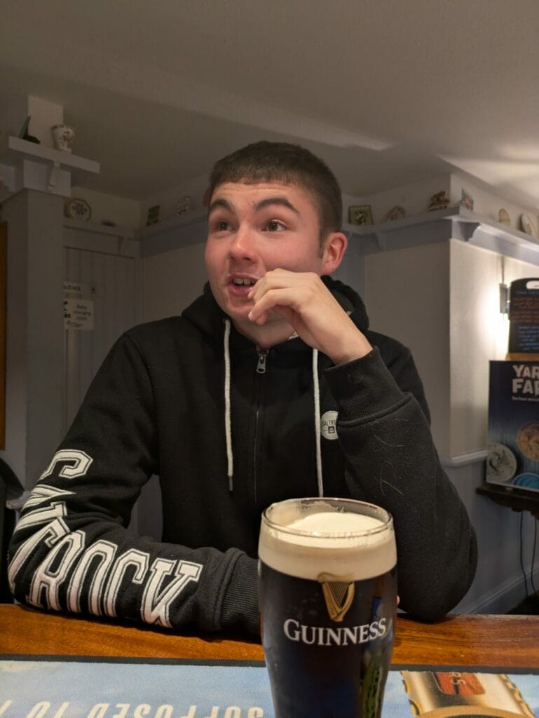 A young man wearing a black hoodie sits at a table indoors, smiling with his hand near his mouth. In front of him is a pint of Guinness. The background shows a decorated wall and shelves.