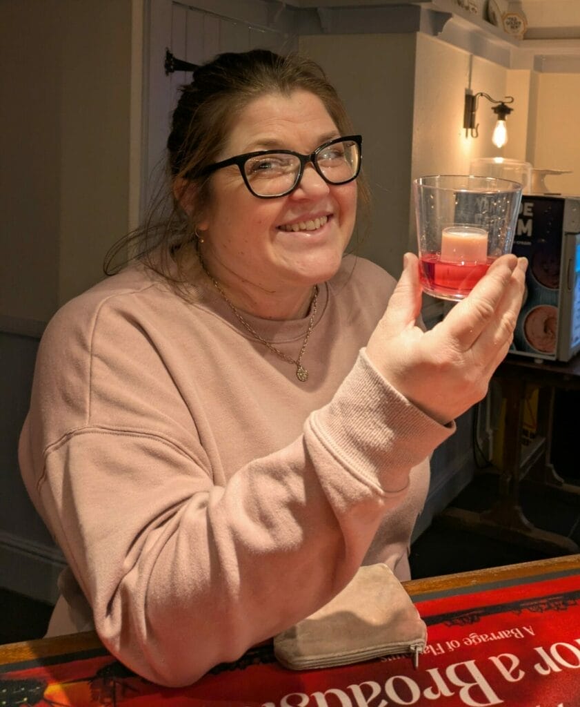 A woman with glasses and a pink jumper smiles while holding up a glass with a red drink and ice, sitting at a bar with a red "Boddingtons" mat in front of her.