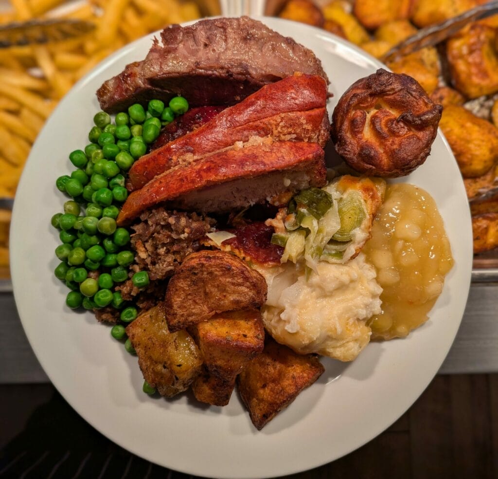A plate filled with roast beef, peas, roast potatoes, stuffing, a Yorkshire pudding, cauliflower cheese, apple sauce, and some glazed carrots, with chips visible in the background. A plate filled with roast beef, peas, roast potatoes, stuffing, a Yorkshire pudding, cauliflower cheese, apple sauce, and some glazed carrots, with chips visible in the background.
